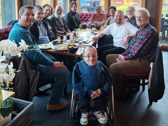 EN in 2nd half – Um einen hellen Tisch herum eng nebeneinander 2 Frauen und 9 Männer in Freizeitkleidung, fünf von ihnen mit Bärten. Sie lächeln in die Kamera. Im Vordergrund der Runde ist ein Mann mit gefalteten Händen in einem kleinen Rollstuhl. Auf dem Tisch vereinzelt Teller und Flaschen und viele Trinkgläser. Im Hintergrund ist die Gruppe von vielen Fenstern umgeben, durch die nachmittägliches Tageslicht zu sehen ist. ENGLISH Two women and nine men in casual clothing are sitting around a bright table, five of them are bearded. They are smiling into the camera. A man with folded hands in a small wheelchair is in the foreground. A small number of plates and bottles and many drinking glasses are on the table. In the background, the group is surrounded by many windows allowing to see the afternoon daylight.