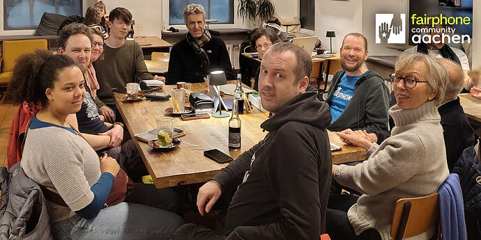 EN in 2nd half – Um einen massiven Holztisch sitzen 4 Frauen und 6 Männer unterschiedlichen Alters in Freizeitkleidung, meist Pullover, und schauen in die Kamera. Drei von ihnen tragen Brillen. Ein Mann im Kapuzenpullover in der Mitte im Vordergrund hat sich zur Kamera gedreht. Auf dem Tisch vereinzelt Trinkgläser, kleine Getränkeflaschen und einige Smartphones. Im Hintergrund sitzen Menschen an Zweiertischen. In der rechten oberen Ecke bedeckt das Logo "fairphone community aachen" einen der Menschen im Hintergrund. ENGLISH Four women and six men of various ages in casual clothing, mostly sweaters, are sitting around a massive wood table and are looking into the camera. Three of them are wearing glasses. A man in a hoodie in the central foreground has turned around towards the camera. A few drinking glasses as well as drinking bottles and a few smartphones are on the desk. In the bacground people are sitting on smaller desks for two. In the top right corner, the logo "fairphone community aachen" covers one of the people in the background.