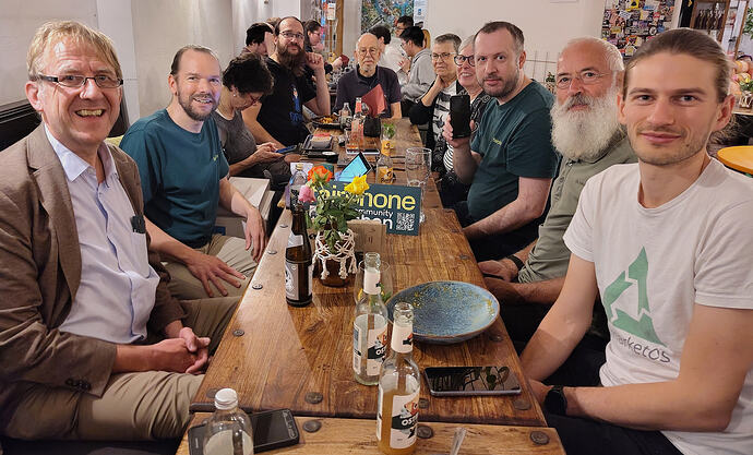 Stammtisch am 25. September 2026 Meetup EN in 2nd half – An einem Tisch, der auf die Kamera zuläuft und auf dem mehrere Flaschen und Gläser und ein Teller stehen, sitzen Menschen unterschiedlichen Alters – vier links, einer am entfernten Ende und fünf rechts. Drei davon sind Frauen, sieben Männer; einige tragen Brillen, einige tragen Bart. Sowohl auf der linken Seite als auch auf der rechten Seite tragen jeweils ein Mann ein waldgrünes T-Shirt. Der auf der rechten hält ein schwarzes Rechteck in seiner Hand und in Richtung Kamera. Bis auf eine Frau auf der Linken, die auf ihr Smartphone schaut, schauen alle in die Kamera. Der Mann ganz rechts trägt ein weißes T-Shirt mit grünem Dreieck und dem Titelfragment "marketOS". ENGLISH: At a table going towards the camera and which has several bottles and drinking glasses and a plate on it, people of various ages are sitting – four on the left, one at the far end and five on the right. Three of them are women, seven men; some are wearing glasses, some are wearing a beard. One man on each the left side as well as on the right side is wearing a forest green T shirt. The one on the right holds a black rectanble in his hand, towards the camera. Apart from one women on the left looking at her smartphone, all are looking into the camera. The man on the far right is wearing a white T shirt with a green triangle and the partial lettering "marketOS".