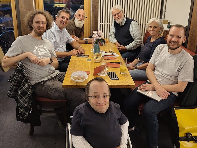 EN in 2nd half – Eine Frau und sechs Männer unterschiedlichen Alters und in Freizeitkleidung, drei von ihnen mit Bart und drei von ihnen mit Brille sitzen um einen Esstisch herum und lächeln in die Kamera. Der Mann im Vordergrund sitzt in einem Rollstuhl. ENGLISH A woman and six men of different ages and in casual wear, three wearing a beard, three wearing glasses are sitting around a dinner table and are smiling into the camera. The man in the foreground is sitting in a wheelchair.