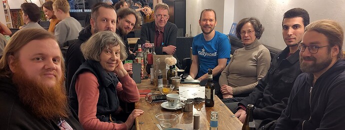 EN in 2nd half – Zehn Menschen (einer verdeckt) verschiedenen Alters sitzen um einen langen Tisch, alle schauen und manche lächeln in die Kamera – drei Männer und eine Frau auf der linken Tischseite, drei Männer und eine Frau. auf der rechten, und ein Man am kurzen Ende des Tisches zwischen ihnen. Im Hintergrund sind noch zwei andere Gruppen, die an anderen Tischen sitzen. ENGLISH: Ten people (one hidden) of various ages are sitting around a long table, all looking and some smiling into the camera – three men and one woman on the left side of the table, three men and one woman on the right, and one man at the small end of the table between them. In the background there are two other groups of people sitting at other tables.