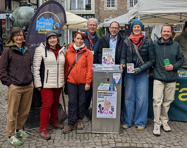 EN in second half – Eine Gruppe von vier Frauen und drei Männern stehen neben und hinter einer silbernen eckigen Tonne, beklebt mit Plakat und Infomaterial mit der Überschrift: "Aachen holt die Handys raus" über dem "Klenkes", einer typisch Aachener Handgeste mit ausgestrecktem kleinen Finger. Hinter der Gruppe ein Aufsteller  mit der Beschriftung "Fairtrade-Stadt Aachen" neben dem blau-grünen Fairtrade-Logo. Alle sieben tragen Jacken und Hosen, feste Schuhe und zwei von ihnen Mützen. Der Mann rechts in grüner Kapuzenjacke hält ein grünes Smartphone. – ENGLISH: A group of four women and three men standing next to and behind a silver edgy bin, a poster and information material sticking on it: "Aachen holt die Handys raus" (Aachen is getting out the mobile phones) above the "Klenkes", a typical local Aachen hand gesture with an erected little finger. Behind the group a stand-up display with the text "Fairtrade-Stadt Aachen" (Faitrade Town Aachen) next to the blue and green Fairtrade logo. All seven are wearing jackets and pants, solid shoes and two of them caps. The man on the right in a green hoodie jacket is holding a green smartphone.