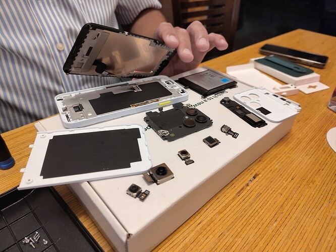 EN in 2nd half – Etwa ein Dutzend weiße und schwarze Einzelteile eines zerlegten Smartphones liegen auf einem kleinen weißen Karton auf einer hölzernen Tischplatte, daneben weiterer Zubehörteile. Hinter dem Tisch ist der untere Teile eines weiß und blau längsgestreiften Hemdes zu sehen und eine Hand, die den abmontierten Bildschirm über den Smartphoneteilen hält. ENGLISH Around a dozen white and black parts of a disassembled smartphone lie on a small white carton box on a wooden desktop, some accessories on the sides. Behind the desk the lower part of a white and blue verticalle striped shirt can be seen as well as a hand that holds the demounted screen above the smartphone parts.