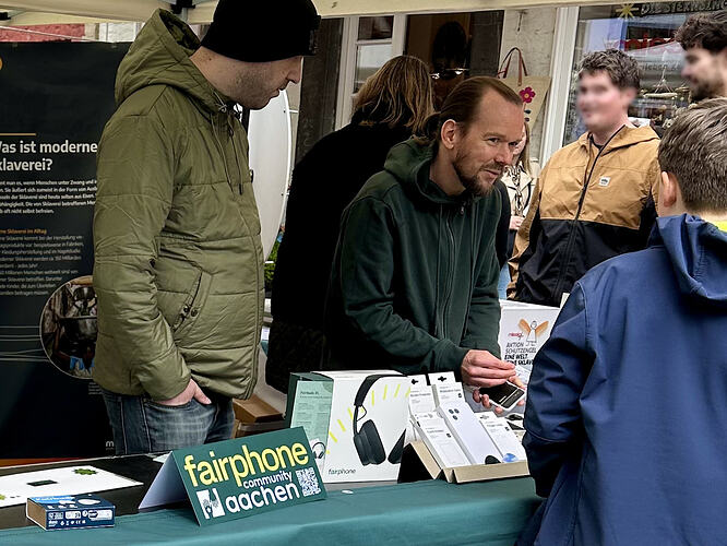 EN in second half – Hinter einem grünen Tisch stehen zwei Männer mittleren Alters in Jacken unterschiedlicher Grüntöne. Der Mann links trägt eine schwarze Mütze, hält eine Hand in der Hosentasche und schaut, wie der bärtige Mann rechts neben ihm sich etwas nach vorn beugt und einen Jungen vor ihm auf der anderen Seite des Tisches am rechten Bildrand anschaut und ihn anzusprechen scheint.. Der Bärtige hält einen schwarzen Gegenstand mit weißer Umrandung in der linken Hand. Hinter ihm eine Frau im schwarzen Mantel und zwei Männer, die sich mit ihr zu unterhalten scheinen. Im Vordergrund links ein grünes Schild "fairphone community Aachen" mit einem Logo und einem QR-Code. – ENGLISH: Two middle-aged men in jackets of differing green colours are standing behind a green desk. The man on the left is wearing a black cap, has his hand in the pocket and watches as the bearded man on his right leans forward a bit and seems to address a boy in front of him on the other side of the desk, at the right of the picture. The bearded man holds a black device with white edges. In the background, a woman in a black coat and two men apparently talking with her. In the foreground on the left, a green sign "fairphone community Aachen" with a logo and a QR code.